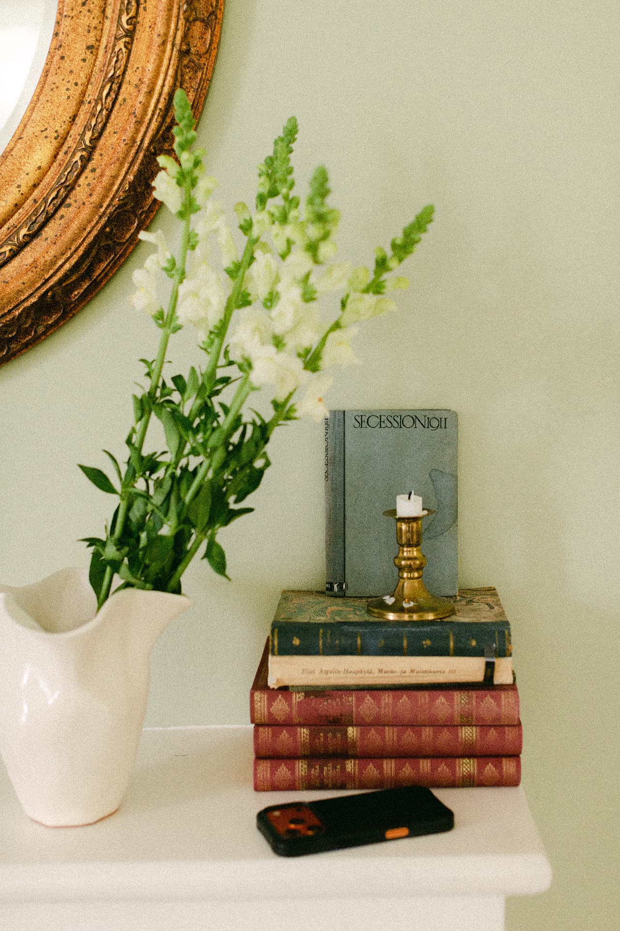 A quiet table with vintage books, a candle, a gold-framed mirror and white flowers